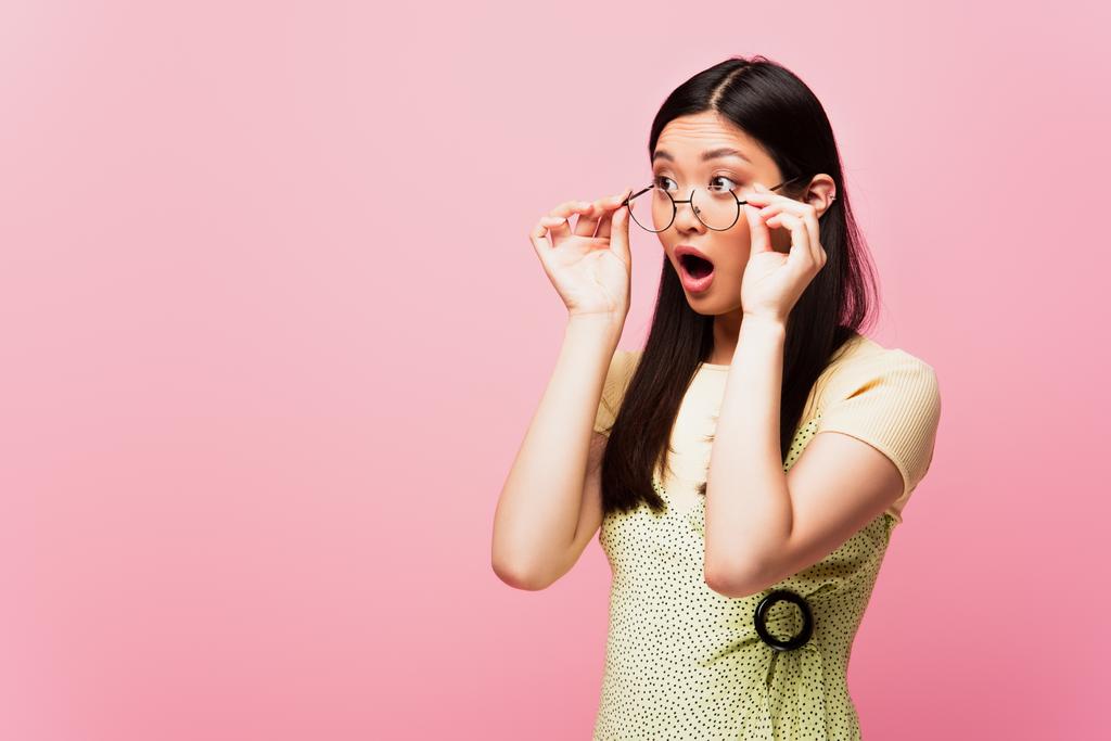 шокированная азиатская девушка с открытым ртом смотрит в сторону и трогательные очки изолированы на розовый  - Фото, изображение
