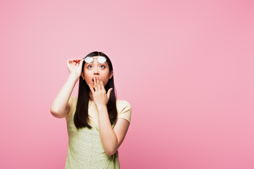 shocked asian girl looking up, covering mouth and touching glasses isolated on pink  - Photo, Image