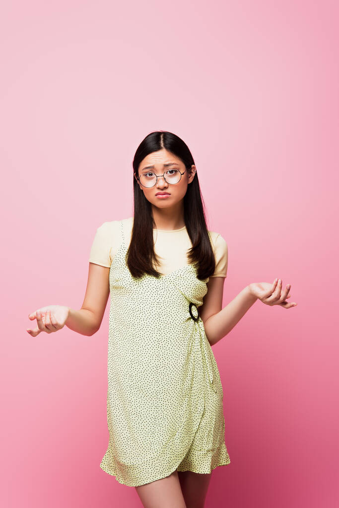 displeased asian girl in glasses showing shrug gesture on pink  - Photo, Image