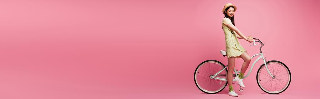 πανοραμική φωτογραφία της χαρούμενης νεαρής Ασιάτισσας με ψάθινο καπέλο κοντά στο ποδήλατο σε ροζ   - Φωτογραφία, εικόνα