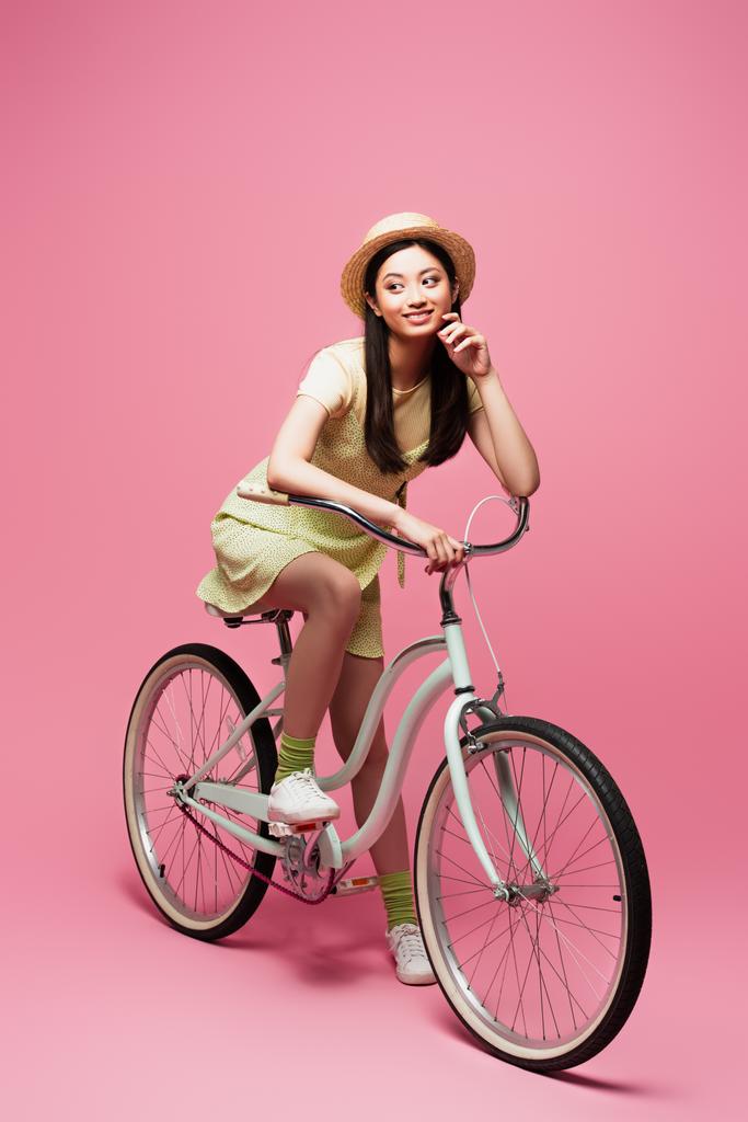 heureux asiatique jeune femme en paille chapeau équitation vélo et regarder loin sur rose   - Photo, image
