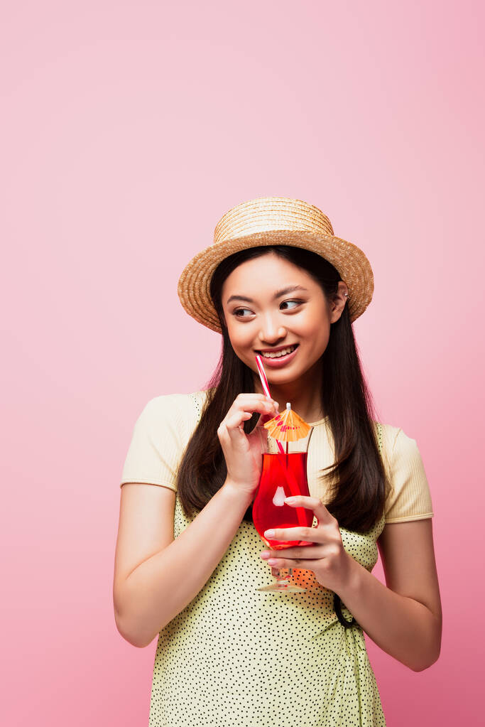 smiling asian woman in straw hat holding glass with cocktail and looking away isolated on pink - Photo, Image