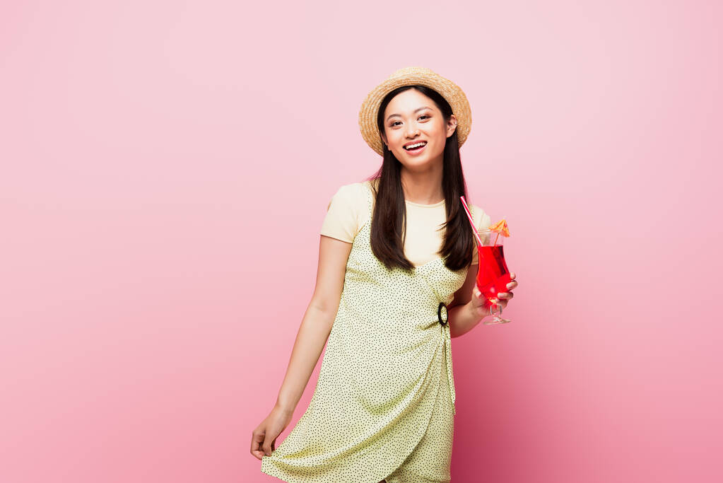 веселая молодая азиатка в соломенной шляпе и платье держа стакан с коктейлем на розовом - Фото, изображение