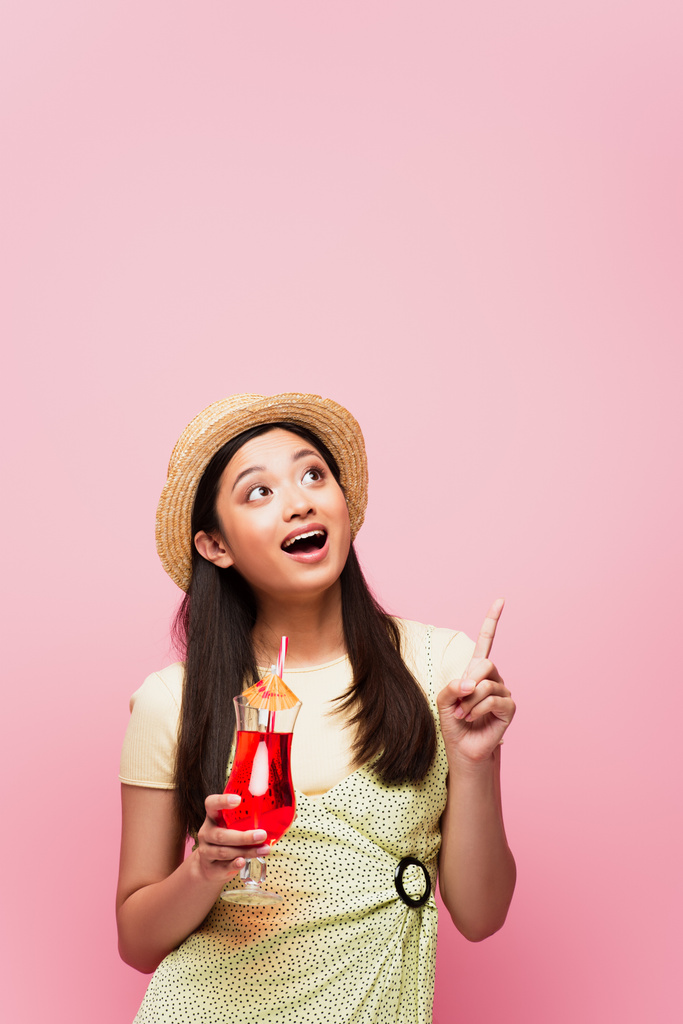 lächelnde junge asiatische Frau mit Strohhut, die Glas mit Cocktail hält und mit dem Finger zeigt, während sie isoliert auf rosa schaut - Foto, Bild
