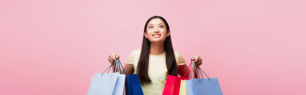imagen horizontal de alegre joven mujer asiática mirando hacia arriba mientras sostiene bolsas de compras aisladas en rosa  - Foto, imagen
