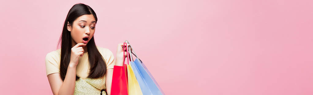 horizontal concept of shocked asian girl looking at shopping bags isolated on pink  - Photo, Image