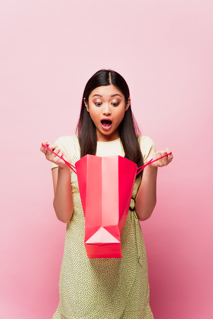 impactado mujer asiática con la boca abierta mirando bolsa de compras en rosa  - Foto, Imagen
