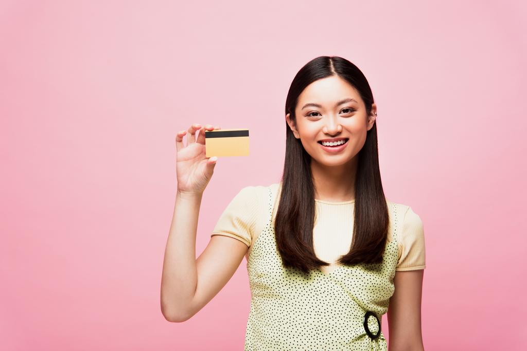 sonriente joven asiática mujer celebración de tarjeta de crédito aislado en rosa  - Foto, Imagen