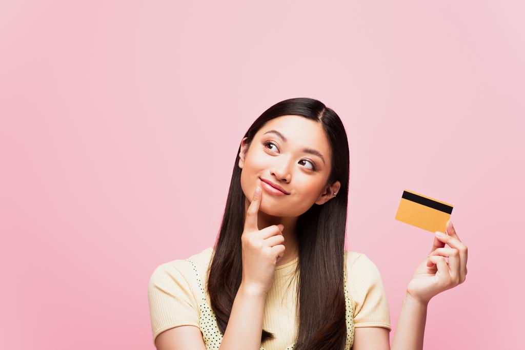 doordachte aziatische vrouw kijken weg en houden credit card geïsoleerd op roze  - Foto, afbeelding