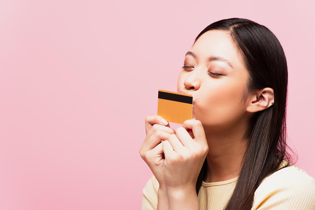 junge asiatische Frau mit geschlossenen Augen küsst Kreditkarte isoliert auf rosa  - Foto, Bild