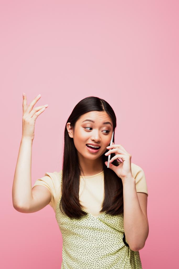 felice donna asiatica gesticolando e parlando su smartphone isolato su rosa  - Foto, immagini