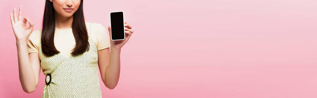 panoramiczny plon młodej kobiety pokazuje znak ok trzymając smartfon z pustym ekranem odizolowany na różowy  - Zdjęcie, obraz
