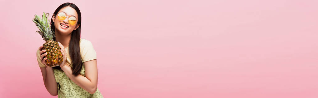coltura orizzontale di felice ragazza asiatica in occhiali da sole tenendo ananas maturo isolato su rosa  - Foto, immagini