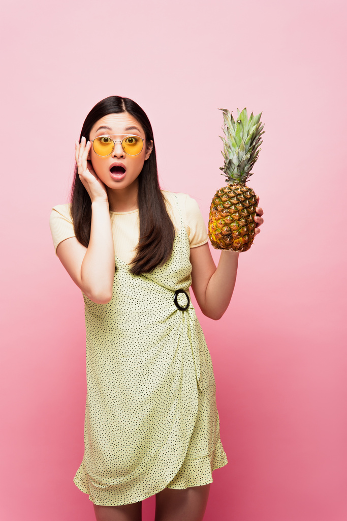 surpris asiatique fille dans lunettes de soleil tenant fraîche ananas sur rose  - Photo, image
