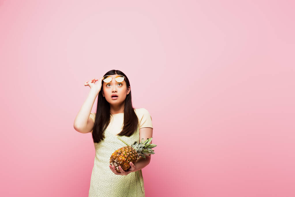 sorpreso asiatico donna in occhiali da sole guardando su e tenendo fresco ananas isolato su rosa  - Foto, immagini