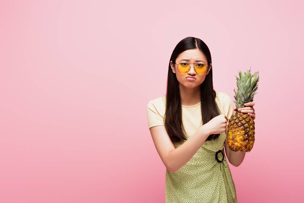 sad asian woman in sunglasses holding fresh pineapple isolated on pink  - Photo, Image