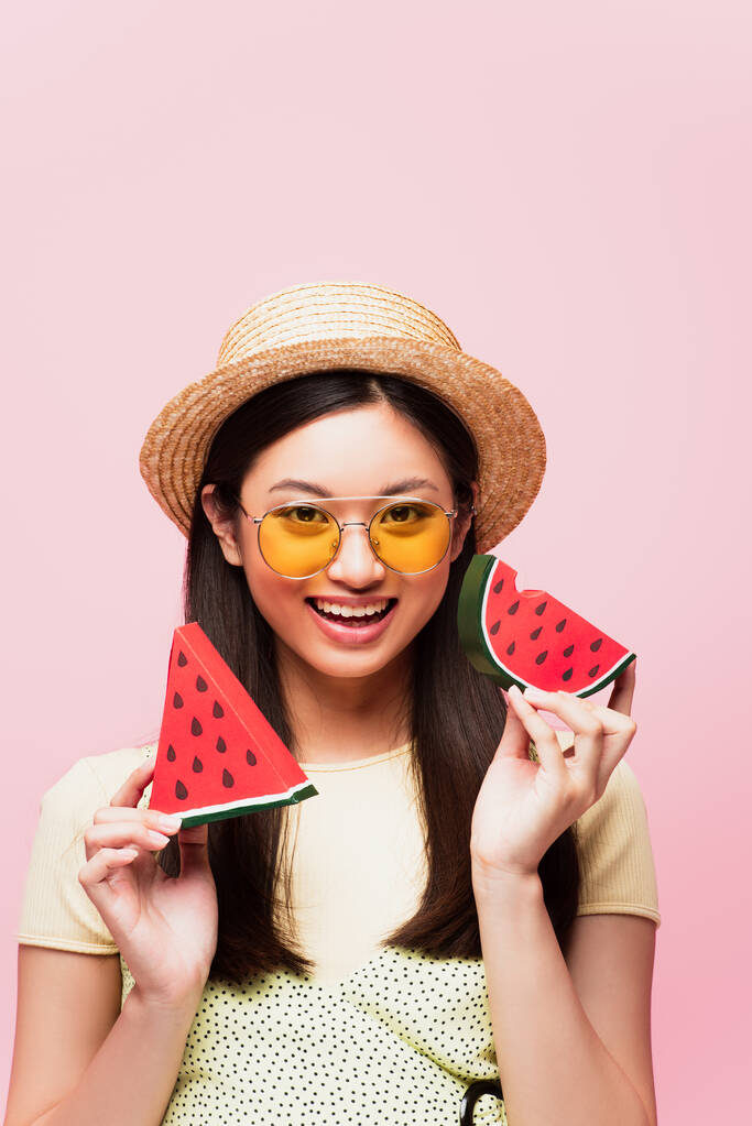 positives asiatisches Mädchen mit Sonnenbrille und Strohhut, das aufgeschnittene Papierwassermelone isoliert auf rosa hält  - Foto, Bild