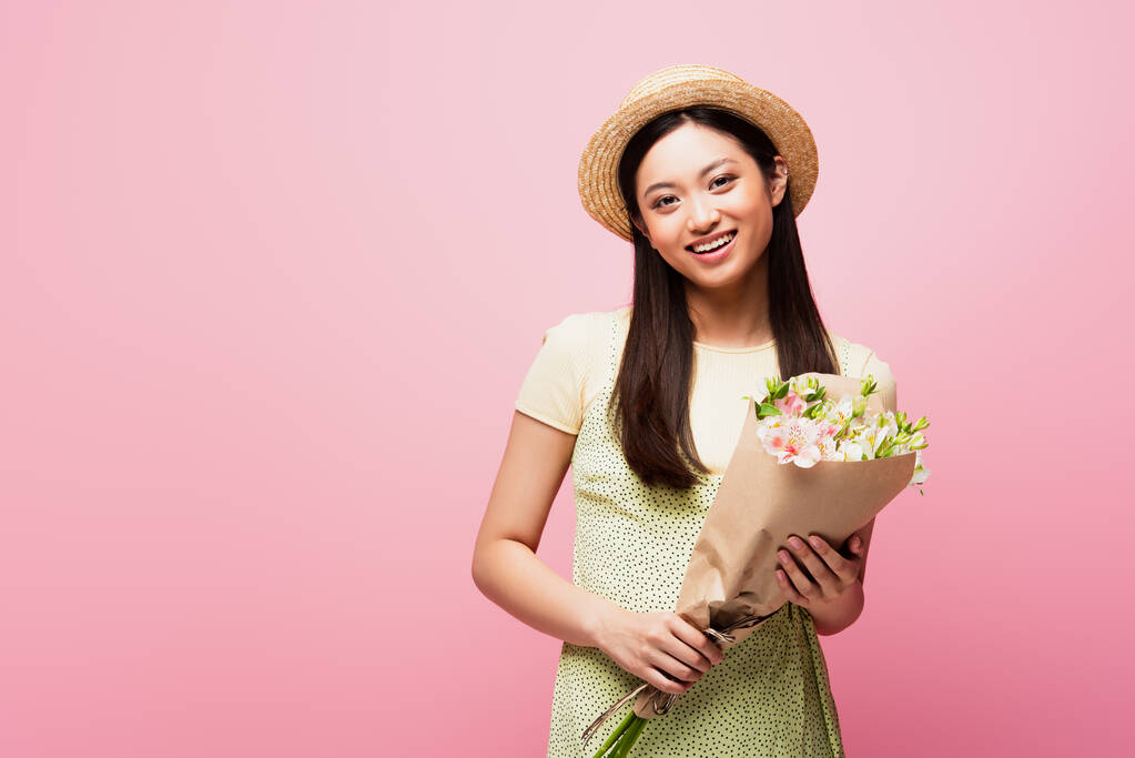 щаслива азіатська дівчина в солом'яному капелюсі тримає букет квітів ізольовано на рожевому
  - Фото, зображення