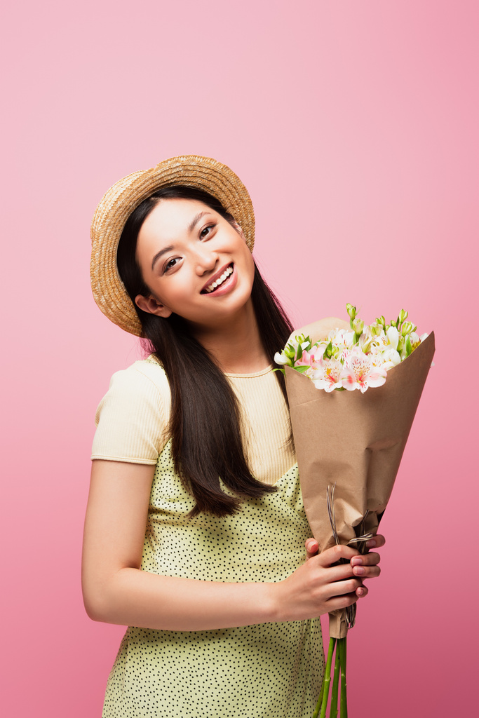 молода азіатка в солом'яному капелюсі посміхається і тримає квіти ізольовані на рожевому
  - Фото, зображення