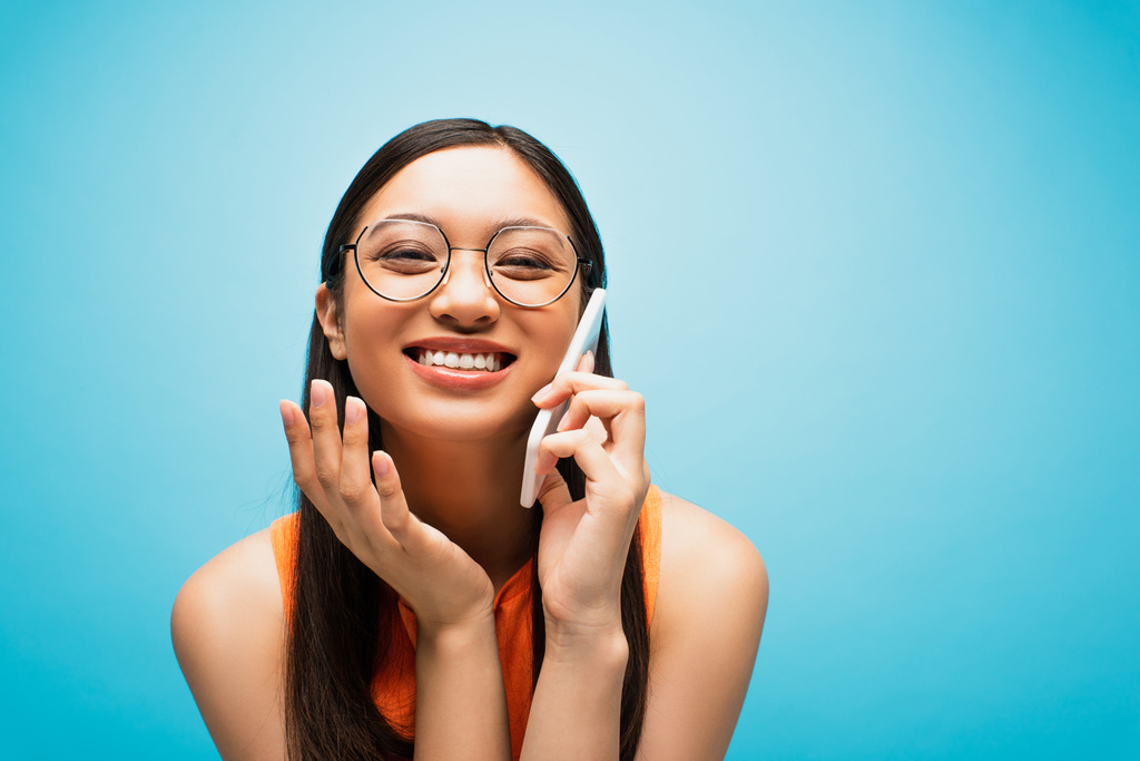 alegre asiático chica en gafas hablando en smartphone en azul - Foto, imagen