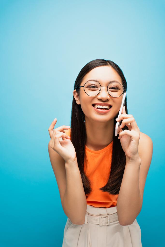 smiling asian girl in glasses talking on smartphone on blue - Photo, Image