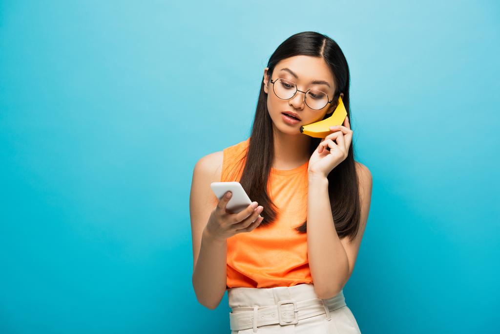chica asiática en gafas usando smartphone y sosteniendo plátano en azul  - Foto, imagen
