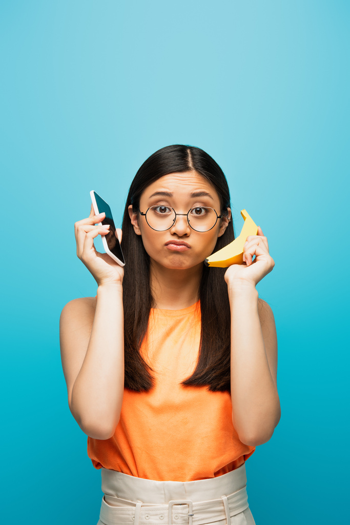 μπερδεμένη Ασιάτισσα με γυαλιά που κρατάει smartphone και μπανάνα απομονωμένη στο μπλε  - Φωτογραφία, εικόνα