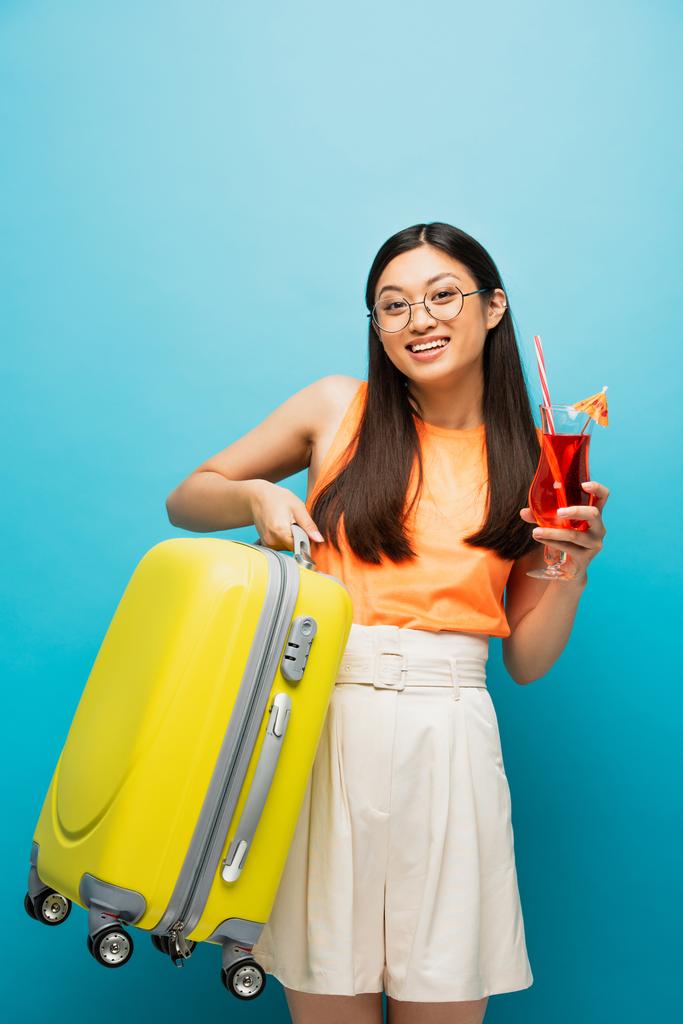 cheerful asian girl in glasses holding cocktail and baggage on blue  - Photo, Image