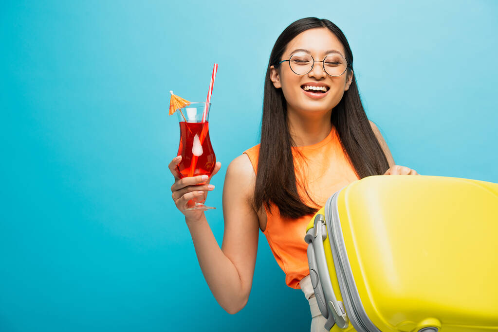 veselá asijská žena v brýlích drží koktejl a zavazadla izolované na modré  - Fotografie, Obrázek