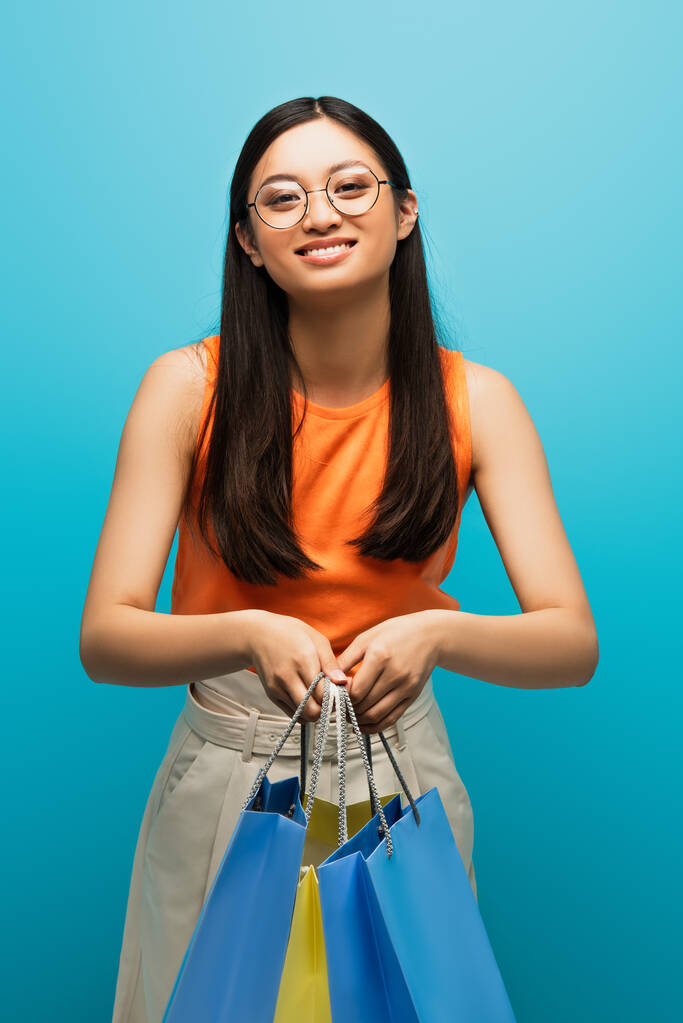 позитивна азіатська жінка в окулярах тримає сумки на синьому
 - Фото, зображення