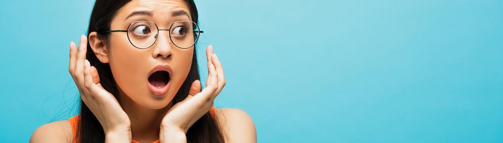 horizontální plodina šokované asijské ženy v brýlích gestikuluje a odvrací pohled izolované na modré - Fotografie, Obrázek