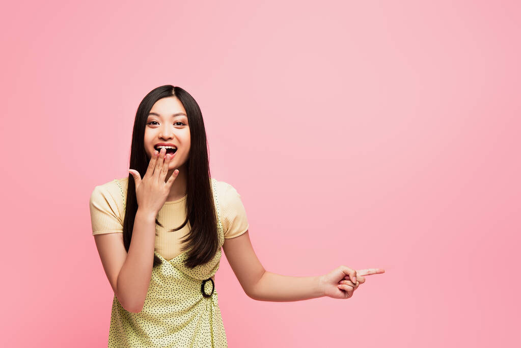 izgatott ázsiai lány ujjal mutogató és fedő száj elszigetelt rózsaszín  - Fotó, kép
