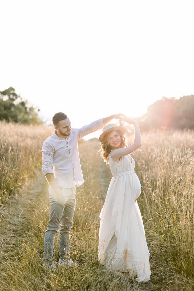 若い白人のカップルは、美しい夏のフィールドで歩くと踊るのが大好きです。帽子と白いドレスの妊婦は彼女のハンサムな男と踊る。幸せな瞬間だ。妊娠の概念は. - 写真・画像