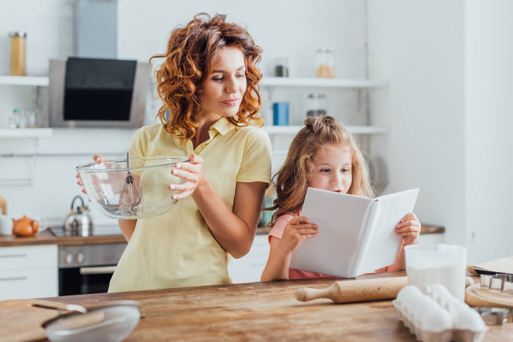 селективное внимание девушки, читающей поваренную книгу рядом с матерью, держащей стеклянную миску с венчиком  - Фото, изображение
