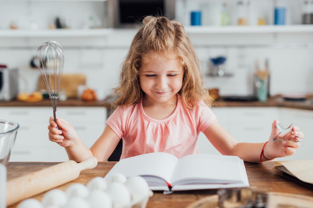 enfoque selectivo de niño sosteniendo batidor mientras lee libro de cocina cerca de huevos de pollo y rodillo - Foto, Imagen