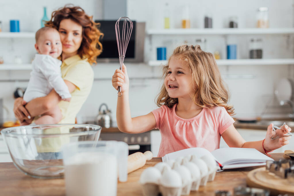 enfoque selectivo del niño sosteniendo batidor cerca de la mesa con ingredientes y utensilios de cocina, y la madre con el bebé en el fondo - Foto, imagen