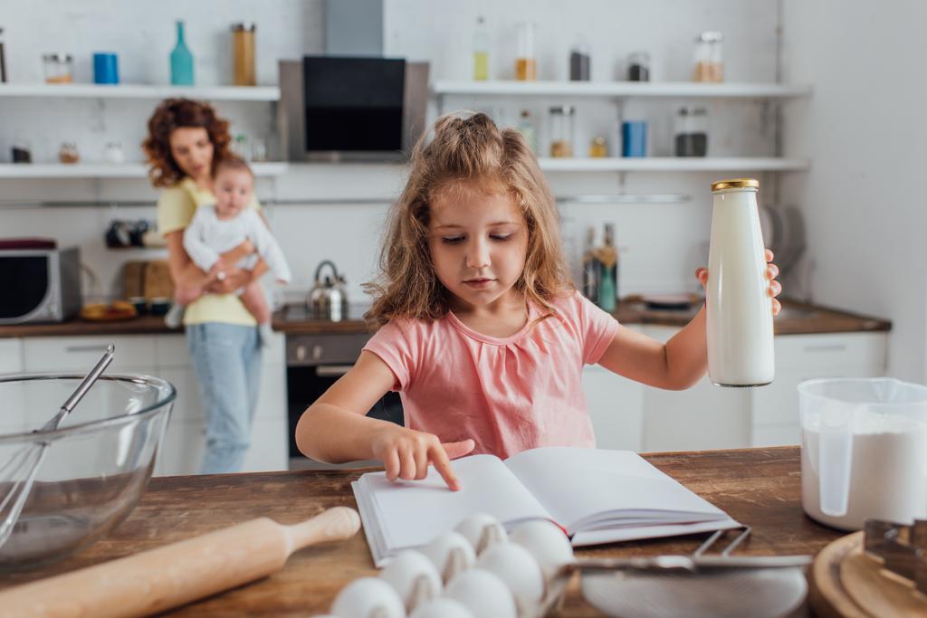 selektivní zaměření dívky drží láhev mléka při čtení kuchařky v blízkosti matky s malým synem - Fotografie, Obrázek