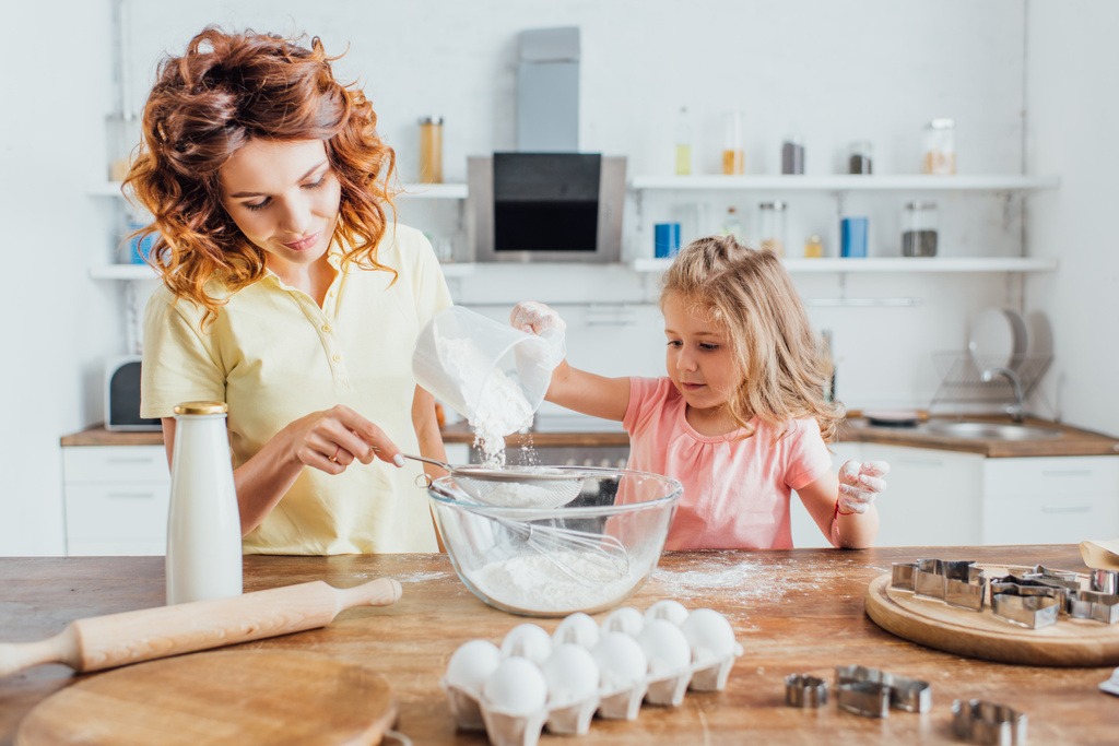 selective focus of young curly woman with daughter sieving flour into glass bowl near ingredients and cookie cutters on table - Photo, Image