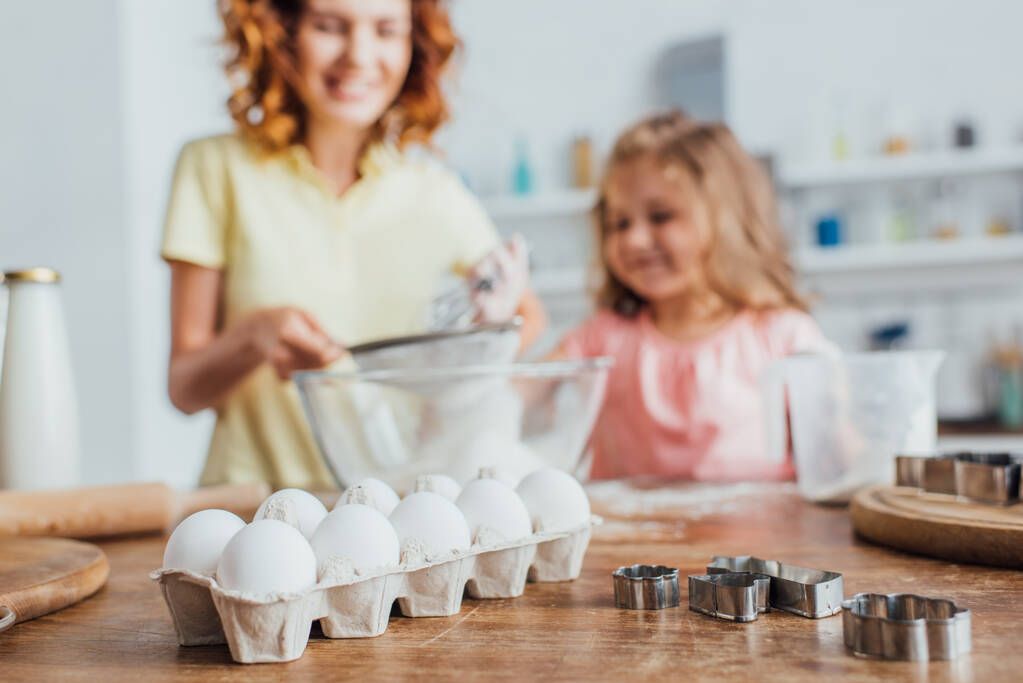 foco seletivo da mulher peneirando farinha em tigela de vidro perto da filha, ovos de galinha na bandeja e cortadores de biscoitos na mesa
 - Foto, Imagem
