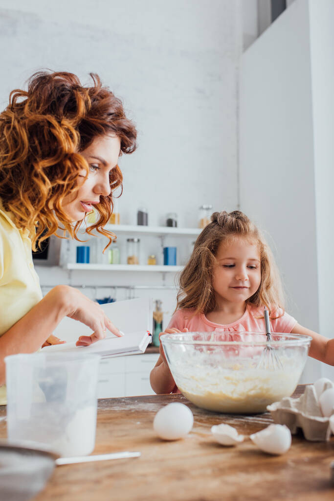 foyer sélectif de maman reding livre de cuisine près de fille et bol en verre avec de la pâte  - Photo, image