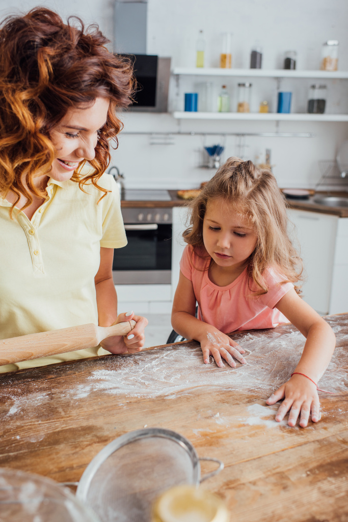 foco seletivo da menina espalhando farinha na mesa da cozinha perto da mãe segurando rolo pin
 - Foto, Imagem