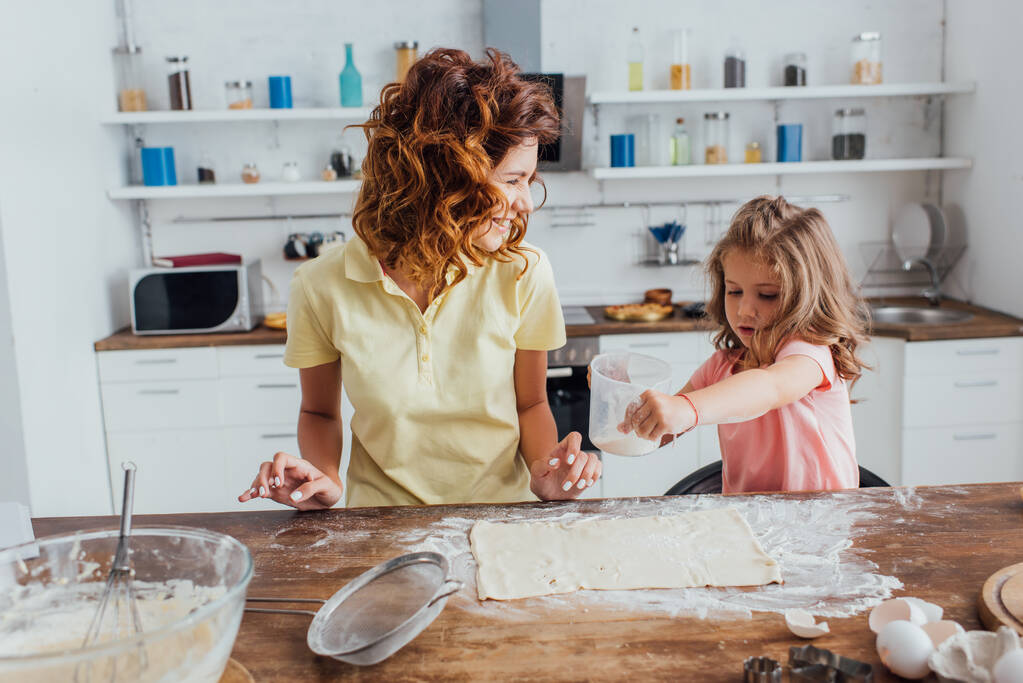 foco seletivo da menina cobrindo massa rolada com farinha perto da mãe na cozinha
 - Foto, Imagem
