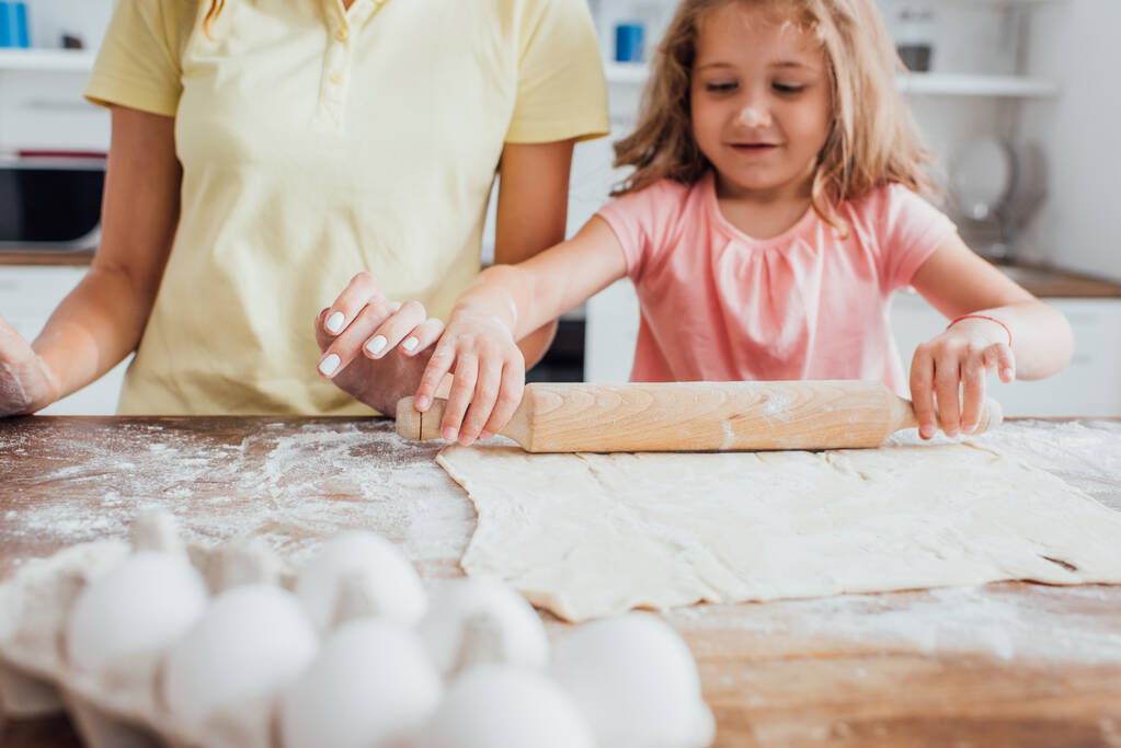 cropped view of mother near daughter rolling out dough on table scattered with flour - Photo, Image