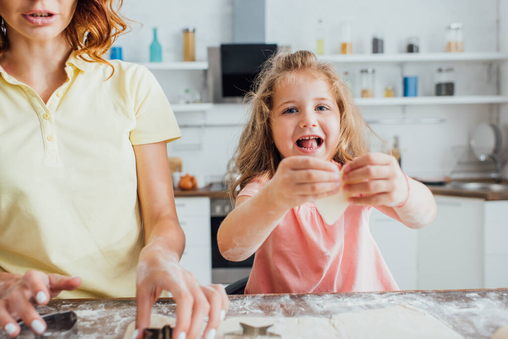selectieve focus van blond meisje met rauw koekje, terwijl moeder snijden gerold deeg met mallen - Foto, afbeelding