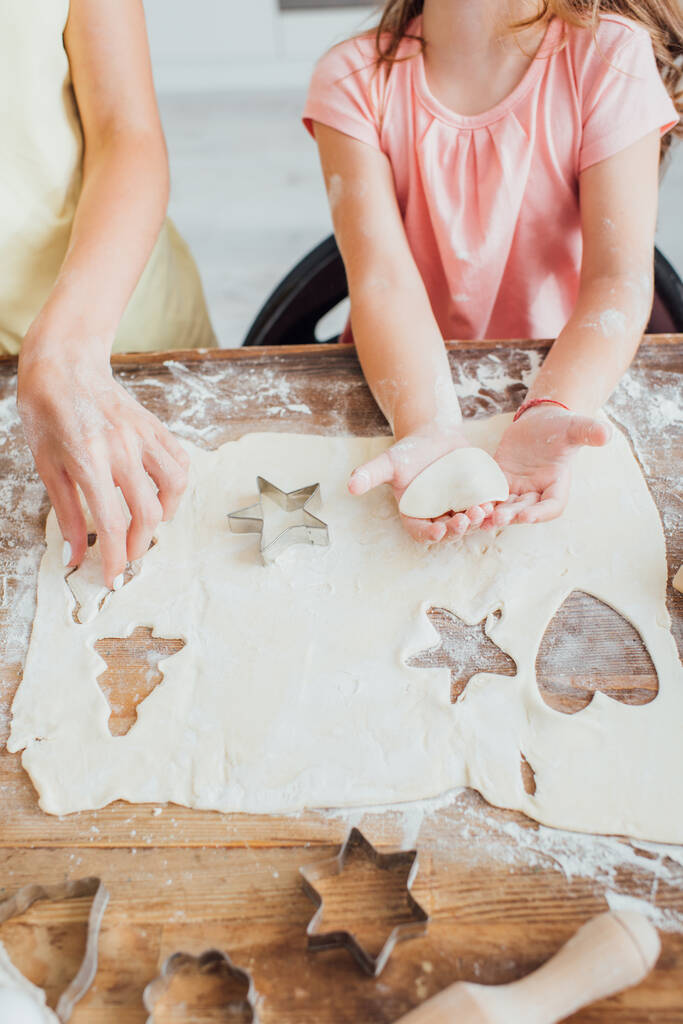 hoge hoek uitzicht van de vrouw met dochter snijden uit multi-vormige koekjes van opgerold deeg op keukentafel - Foto, afbeelding