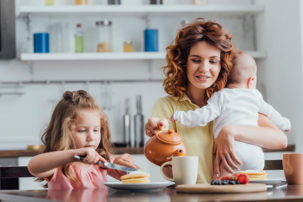 selective focus of mother pouring tea from teapot while holding infant near daughter eating pancakes - Photo, Image
