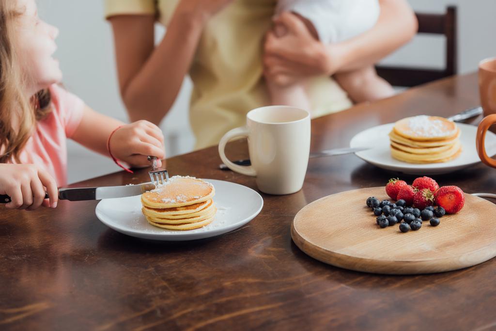 cropped view of mother holding infant son near daughter eating delicious pancakes, selective focus - Photo, Image