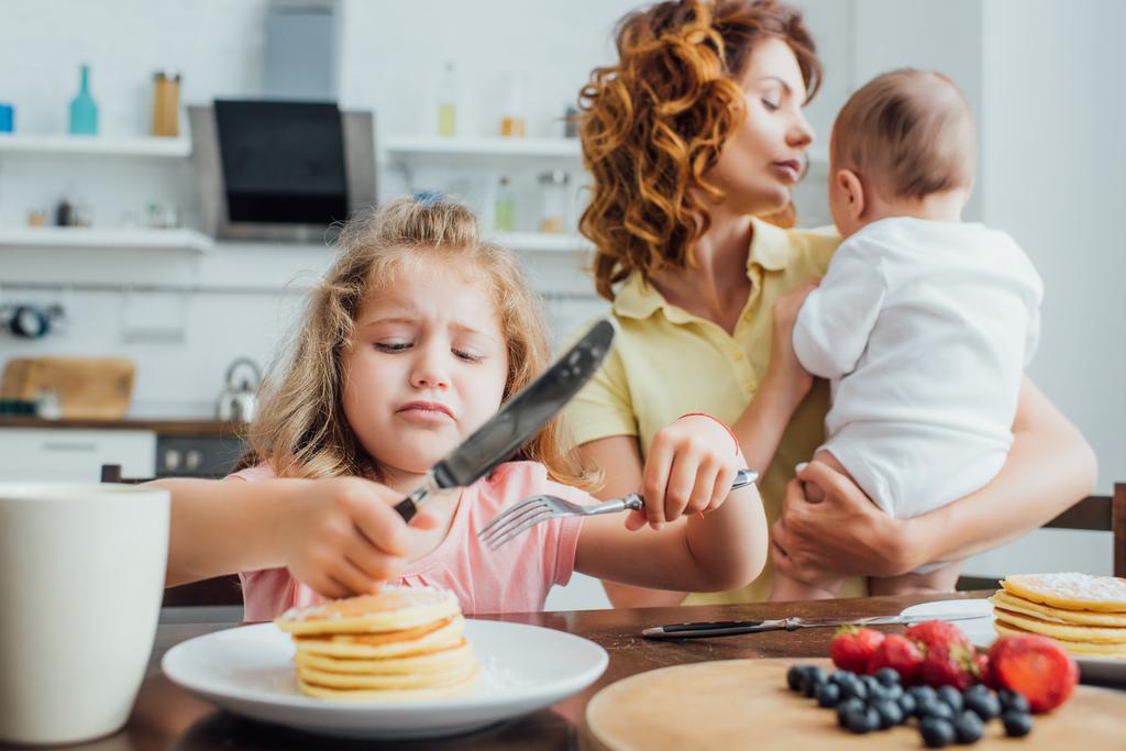 enfoque selectivo de niña disgustada sosteniendo tenedor y cuchillo cerca de panqueques mientras madre sosteniendo niño - Foto, imagen