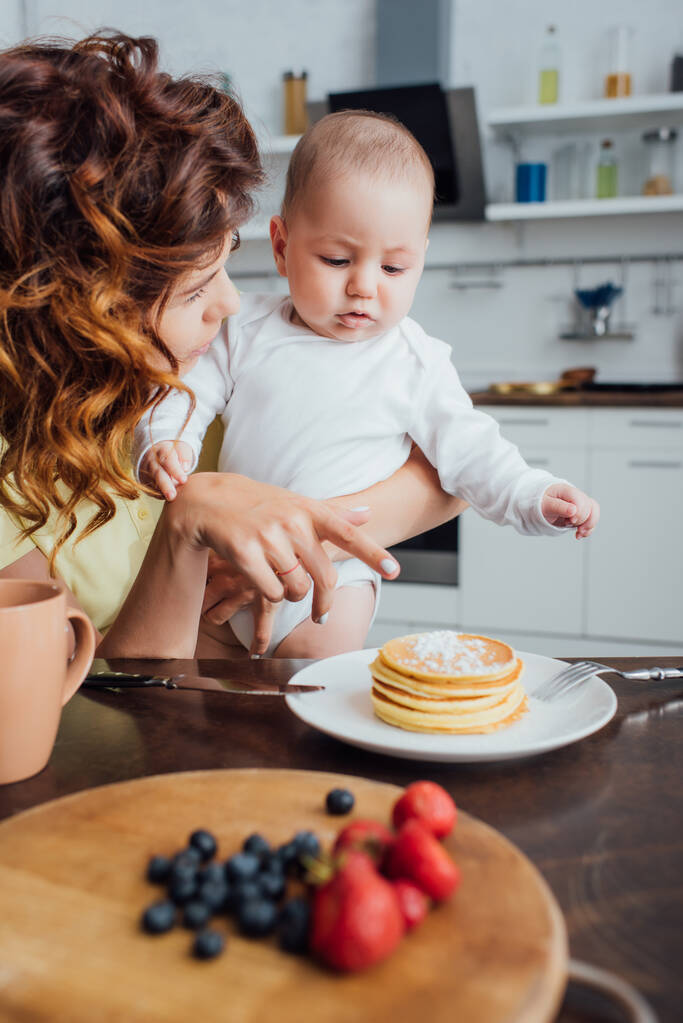 selectieve focus van jonge moeder wijzend met de vinger naar heerlijke pannenkoeken terwijl het houden van zuigeling zoon - Foto, afbeelding