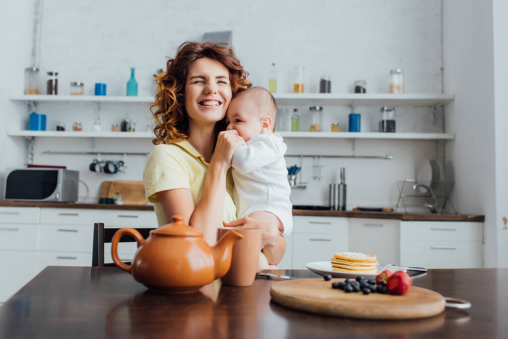 селективное внимание молодой матери, держащей младенца в комбинезонах возле стола, подаваемого с завтраком  - Фото, изображение